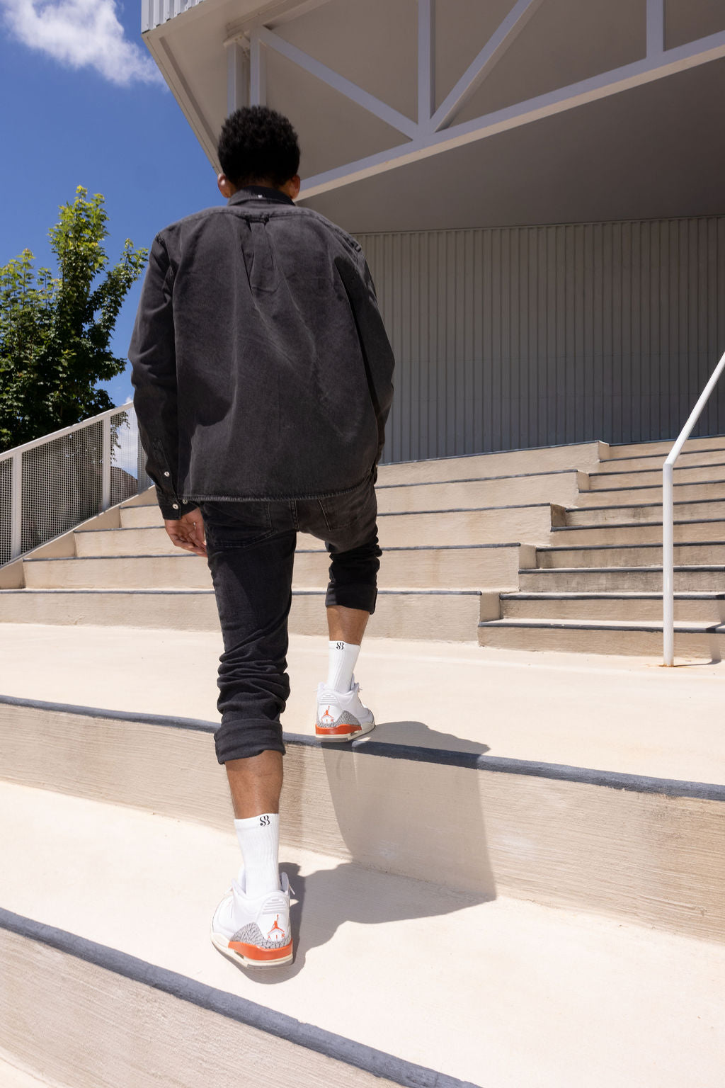 A man wearing a dark gray jacket, black pants, white socks, and white sneakers with orange accents walks down outdoor concrete stairs under a blue sky.