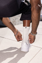 A close-up lifestyle shot of a person tying the laces of a white sneaker, wearing a white ribbed crew sock with a black monogram logo, black shorts, and silver bracelets on a light stone tiled floor.