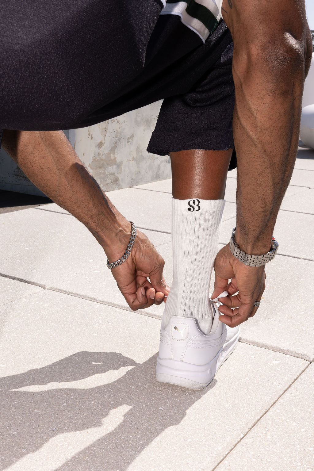 A close-up lifestyle shot of a person tying the laces of a white sneaker, wearing a white ribbed crew sock with a black monogram logo, black shorts, and silver bracelets on a light stone tiled floor.