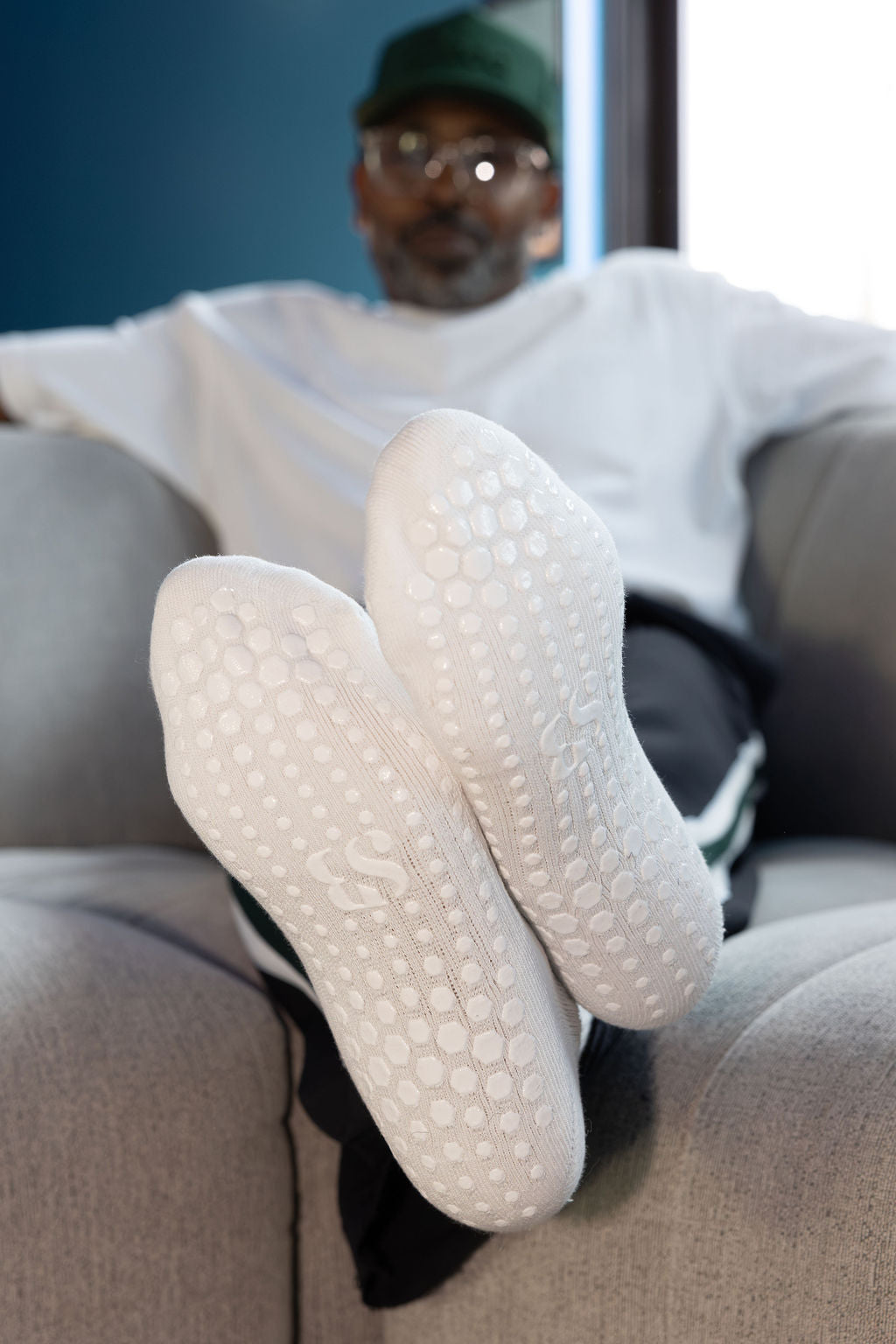 A person reclining on a gray couch with their feet raised, wearing white textured slip-on shoes. The shoes have a raised circular tread pattern and a subtle embossed logo on the sole.