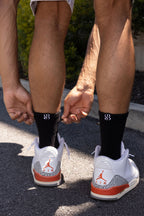 Close-up of a person tying black crew socks with a white and orange Air Jordan sneaker visible on one foot, outdoors on asphalt with greenery in the background.