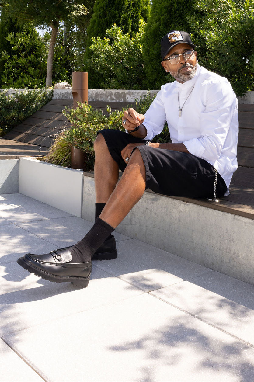 A man wearing a white long-sleeve shirt, black shorts, black loafers, and a black cap sits on a concrete bench in an outdoor garden setting.