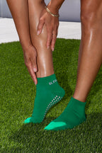 A close-up of a person wearing bright green pointed-toe grip socks on grass, with one hand touching the calf and the other holding the foot. The brand name SLTG is printed in white on the ankle.