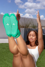 A woman outdoors on grass with her legs raised, wearing bright green textured socks with white dots and white ankle bands. The socks have a visible 'S' logo, and the background shows a modern wall, sky, and clouds.