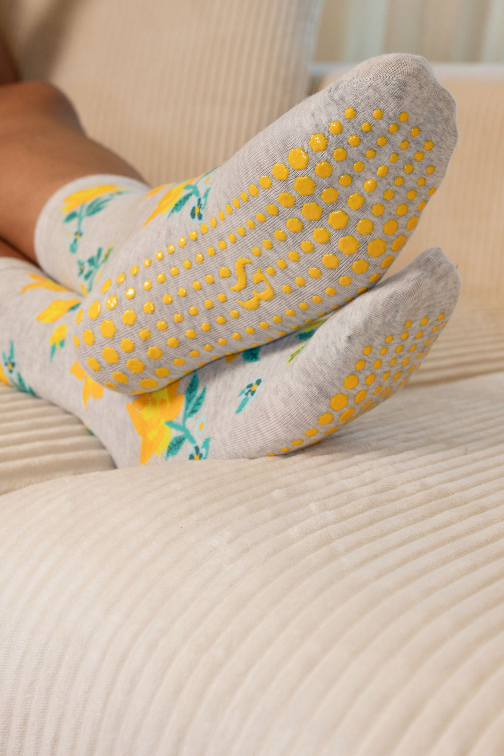 Close-up of a person's feet wearing light gray grip socks with a yellow and green citrus pattern, resting on a ribbed cream-colored couch.